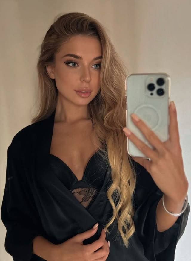 A mirror selfie captures a woman in a black robe over a lace bra, holding a light-colored phone against a plain beige wall.