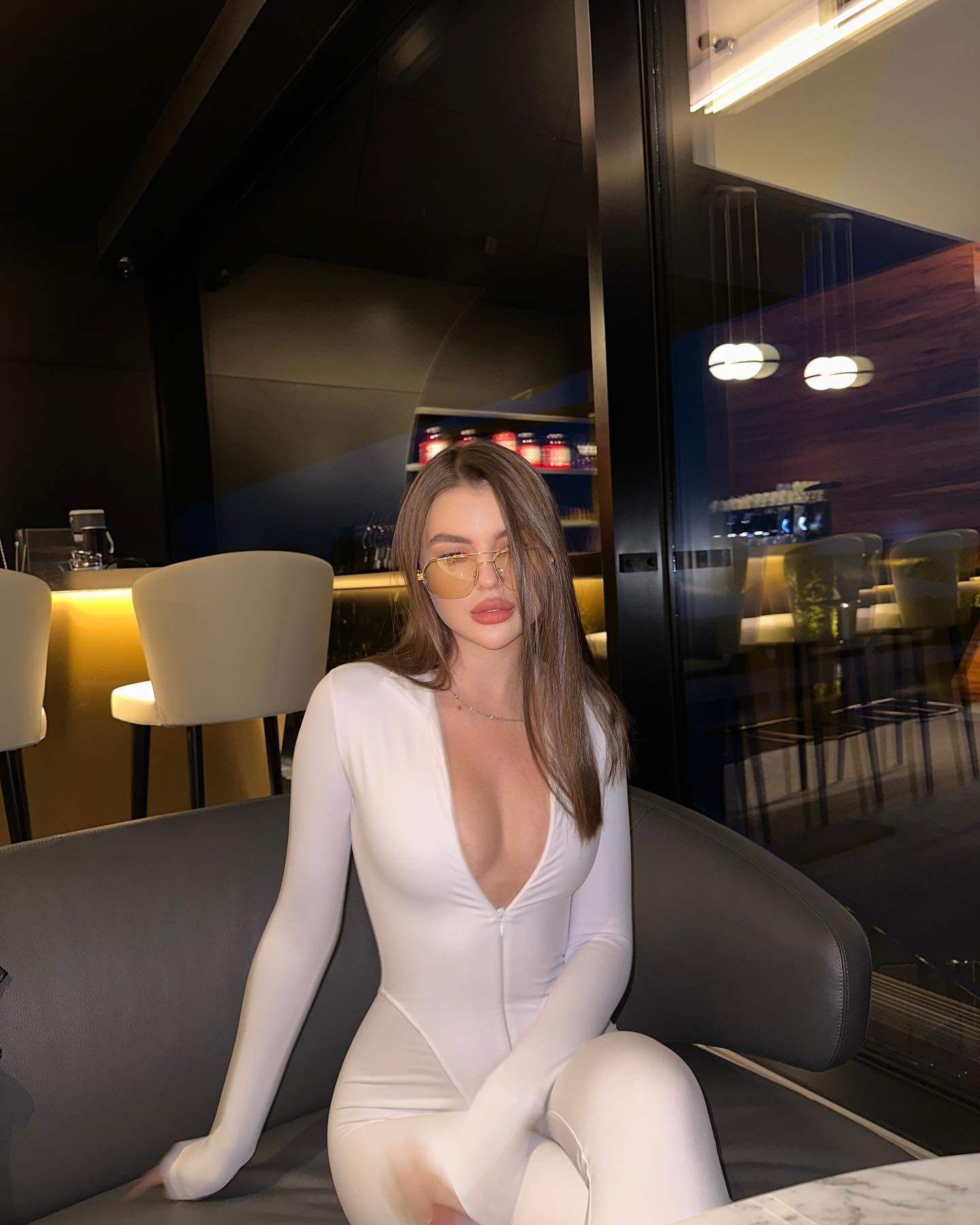 A woman with sunglasses sits on a dark gray sofa in a dim lounge, wearing a white long-sleeved bodysuit with a deep V-neckline and front zipper.