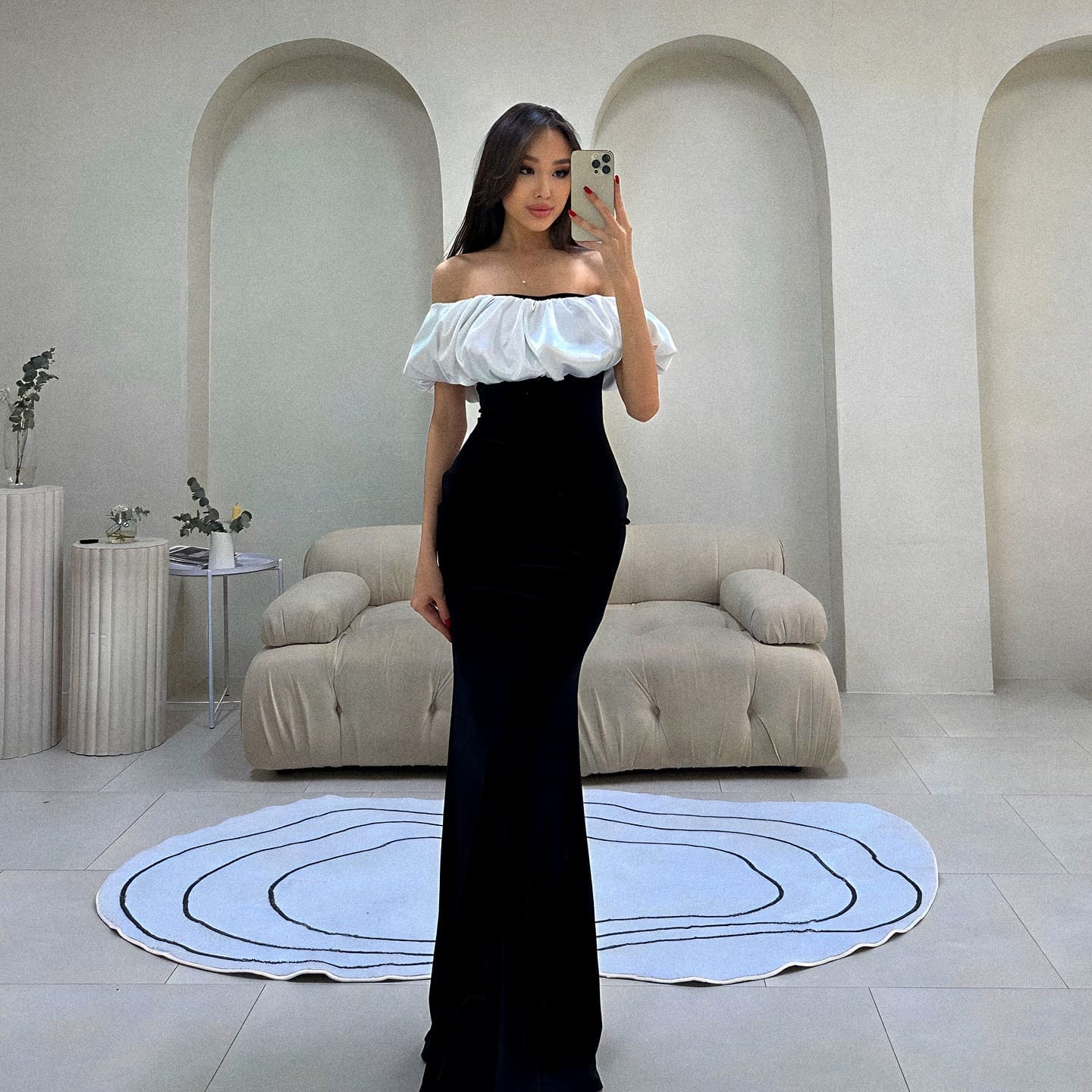 A woman stands in a room with arched niches, wearing a black dress with a white off-the-shoulder top, holding a phone over her face.