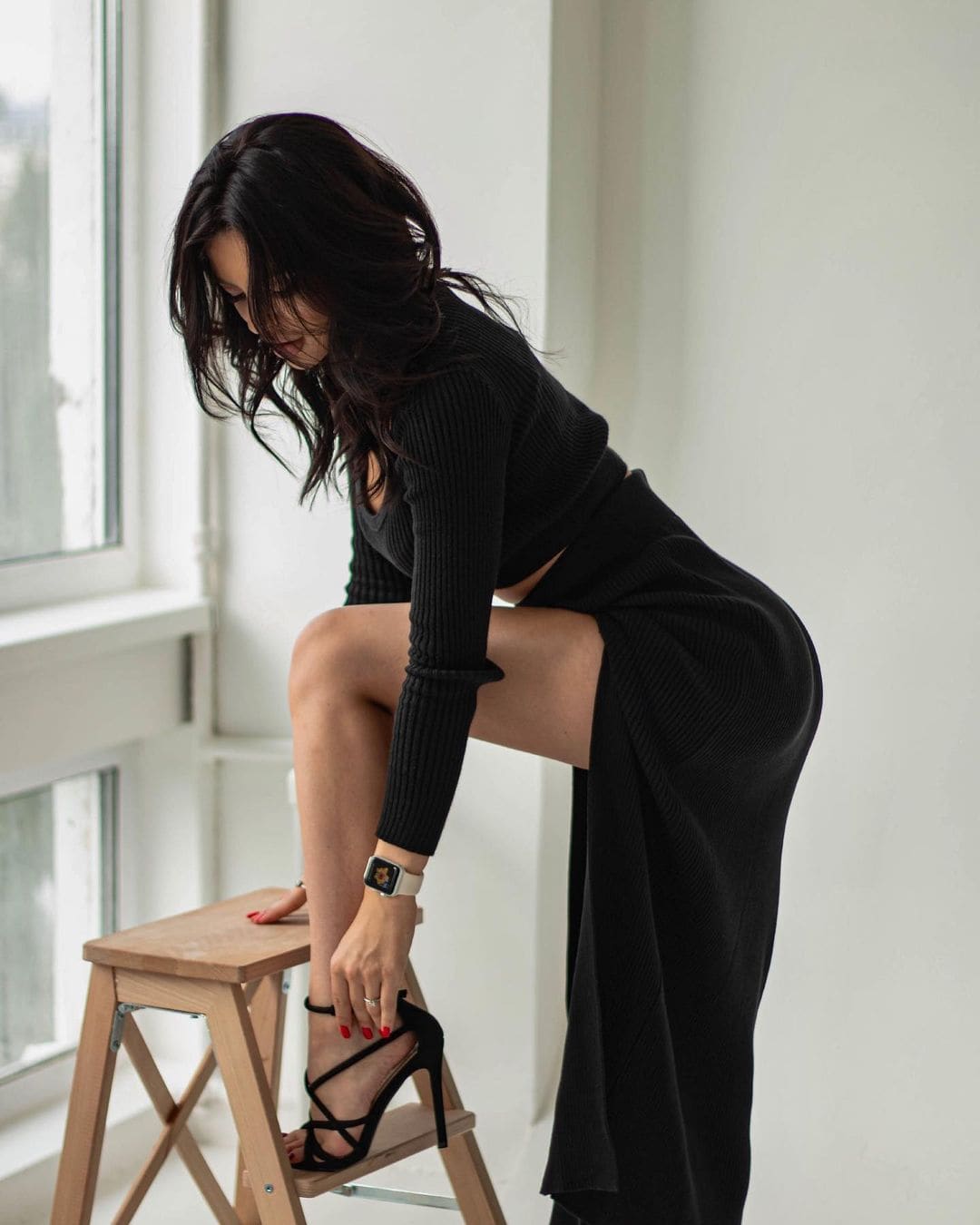 Bending on a wooden stool, a woman in a black top and skirt adjusts a high heel in a bright room.