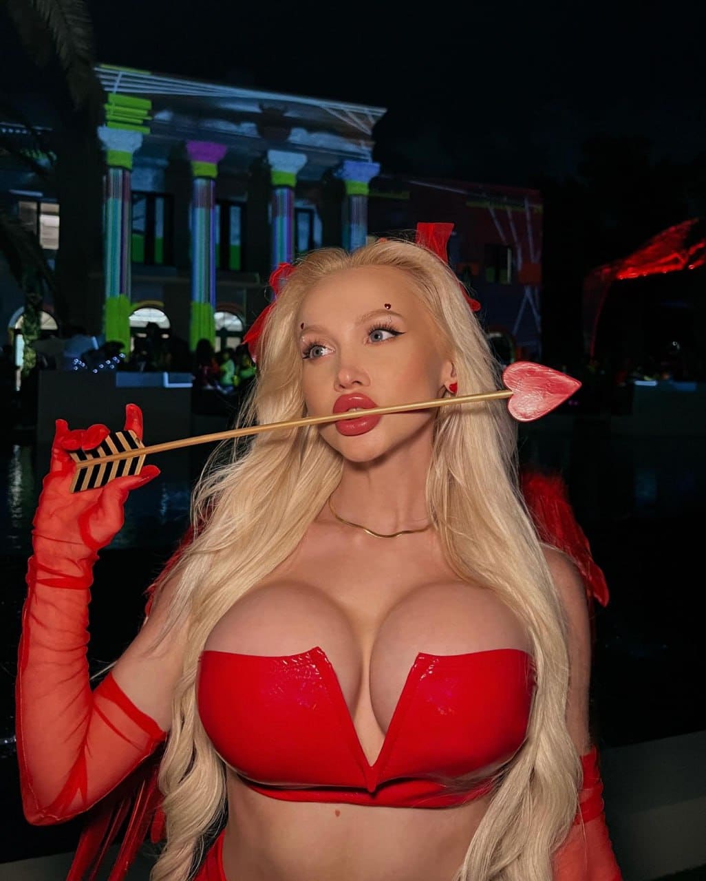 A blonde woman in a red strapless top and sheer gloves holds a prop arrow in her mouth, standing before a building with colorful columns at night.