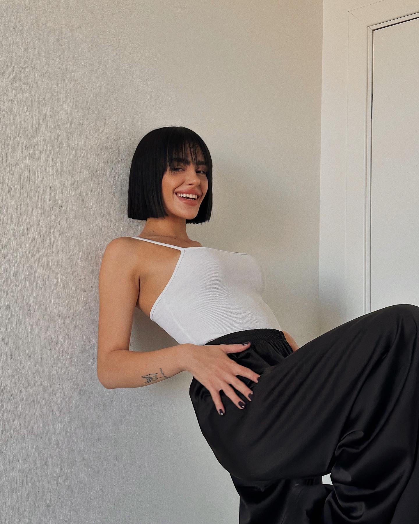 Smiling and leaning on a wall, a woman in a white tank top and black trousers rests a hand on her hip.