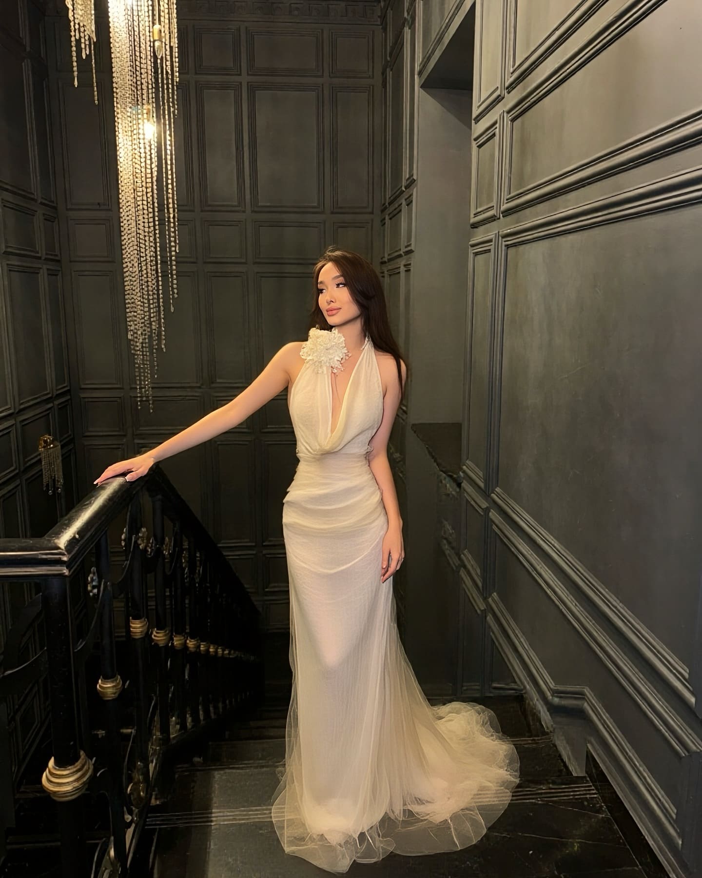 A woman stands on a dark stone staircase in a white halter gown with a deep V-neck and floral collar, near a beaded chandelier.
