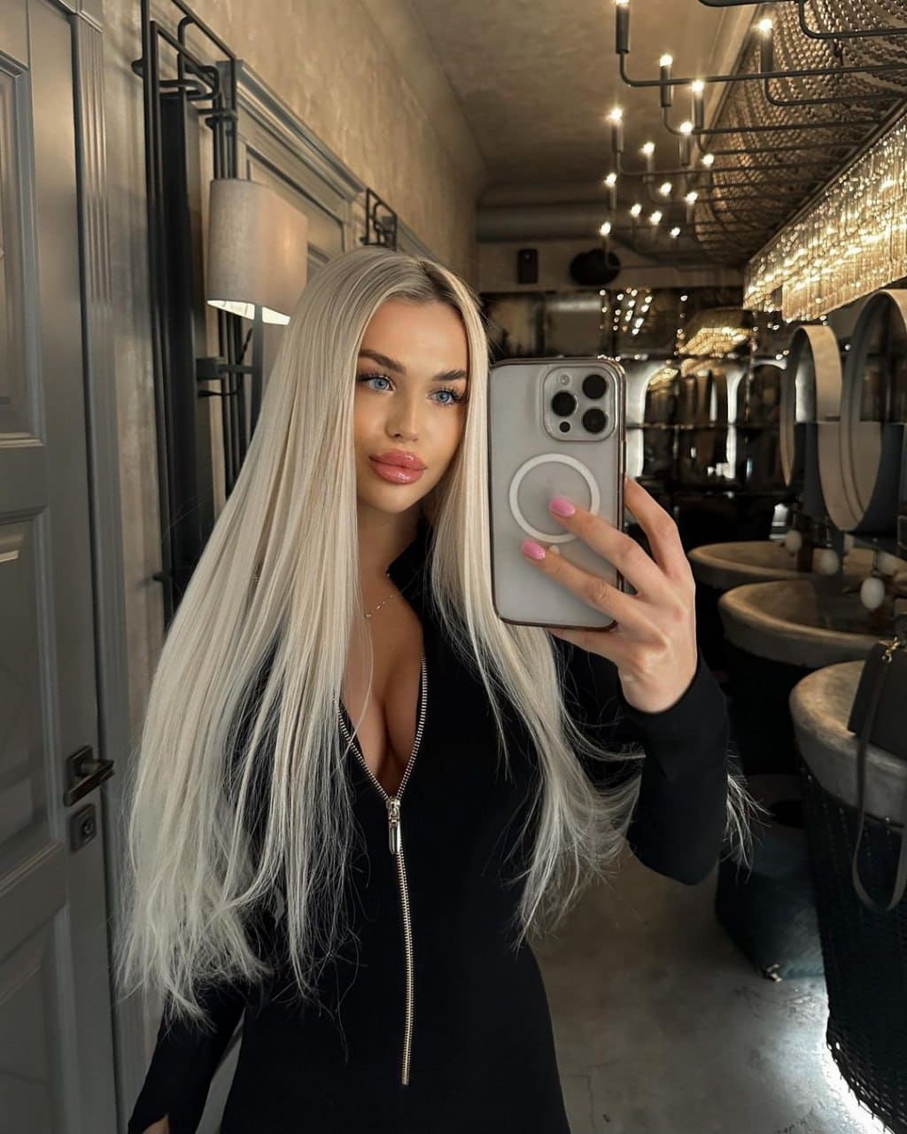 Jasmina takes a mirror selfie in a restroom, wearing a black zip-front top and holding a silver phone.