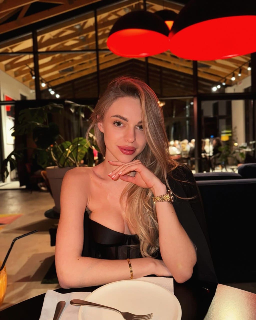 Vanessa sits at a restaurant table under red lights, wearing a black strapless top and resting her chin on her hand.
