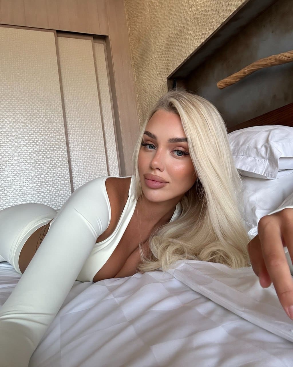 A blonde woman lies on a bed with white sheets, wearing a white long-sleeved top with a deep neckline and matching bottoms near a wooden headboard.