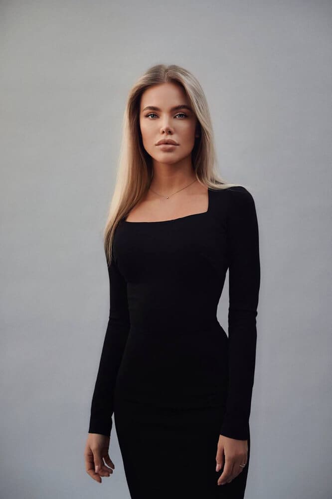 Luiza stands facing forward in a fitted black long-sleeve dress with a square neckline and thin necklace against a gray studio background.