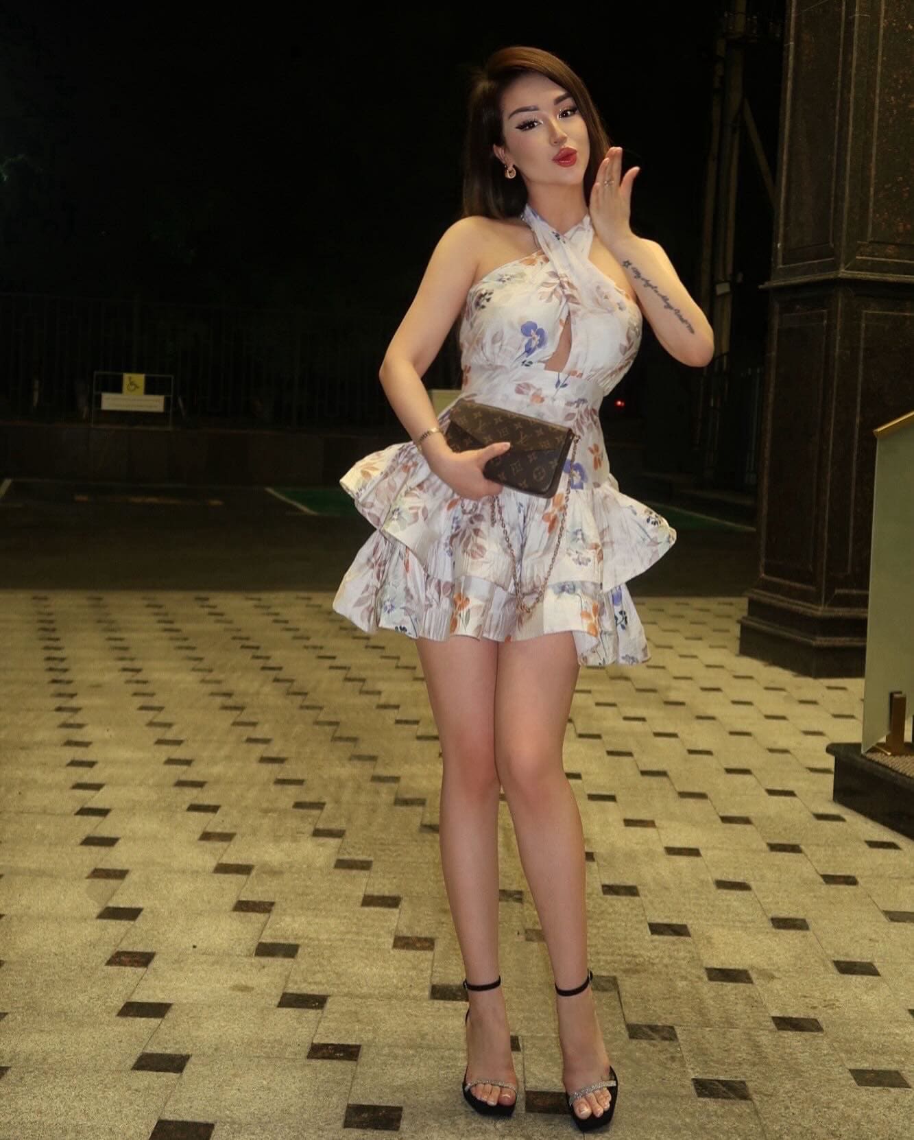 At night on pavement, she wears a white floral halter dress and holds a patterned clutch near her face.