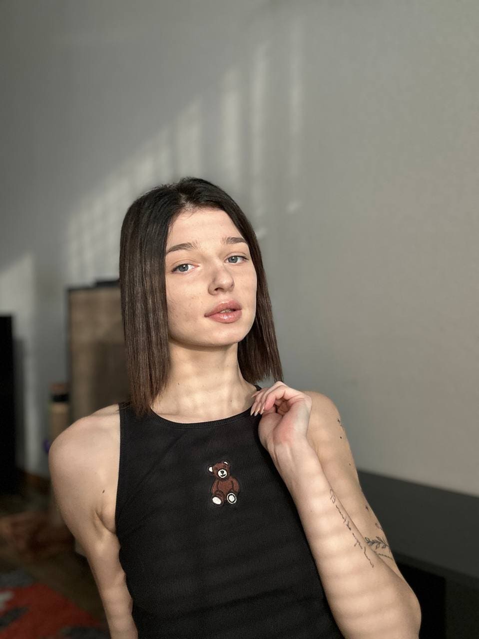 Violet stands indoors against a plain wall, wearing a black tank top with a bear, showing a forearm tattoo.