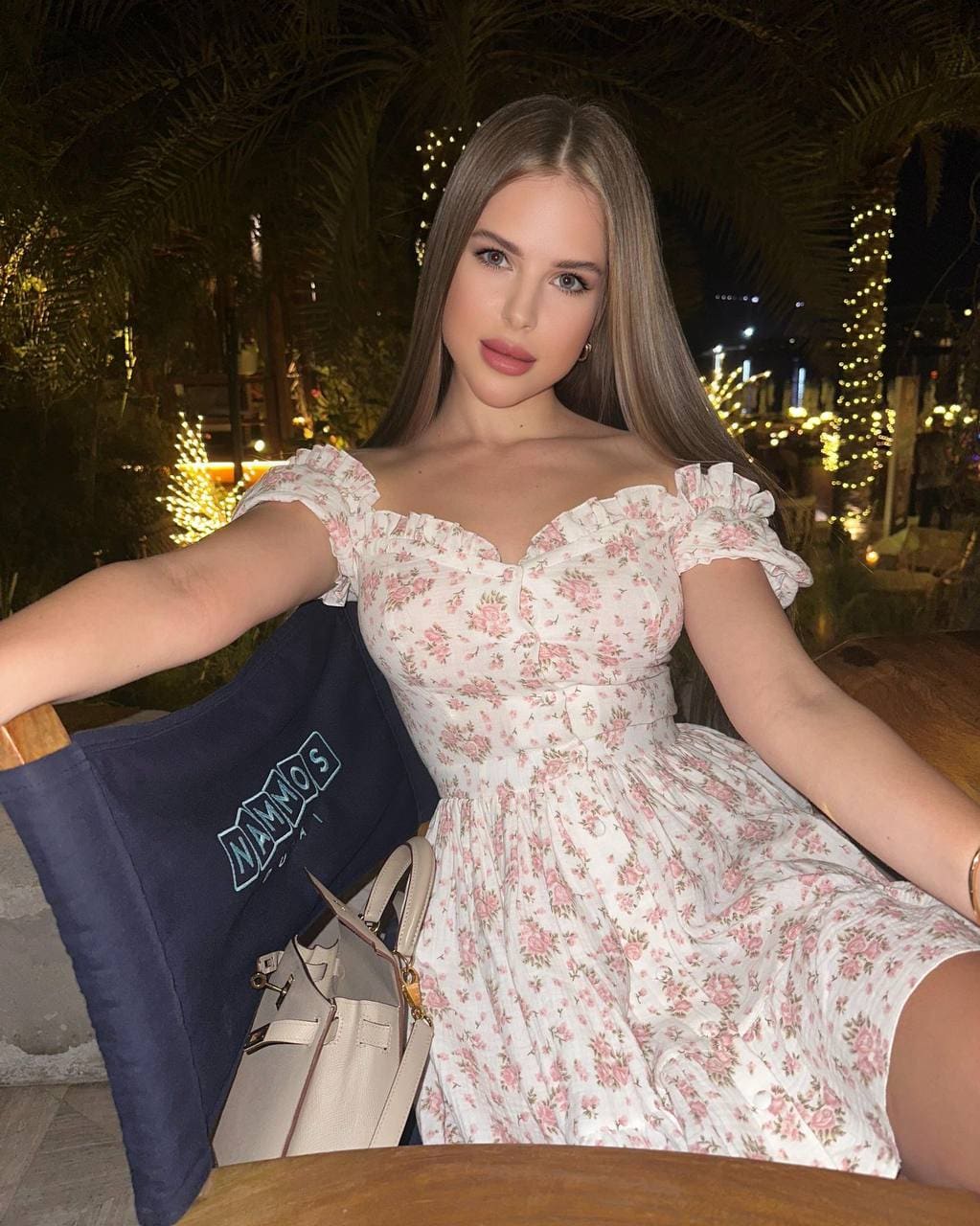 Lyubava sits at a wooden table outdoors at night in a white off-shoulder dress with pink floral print and ruffled sleeves.