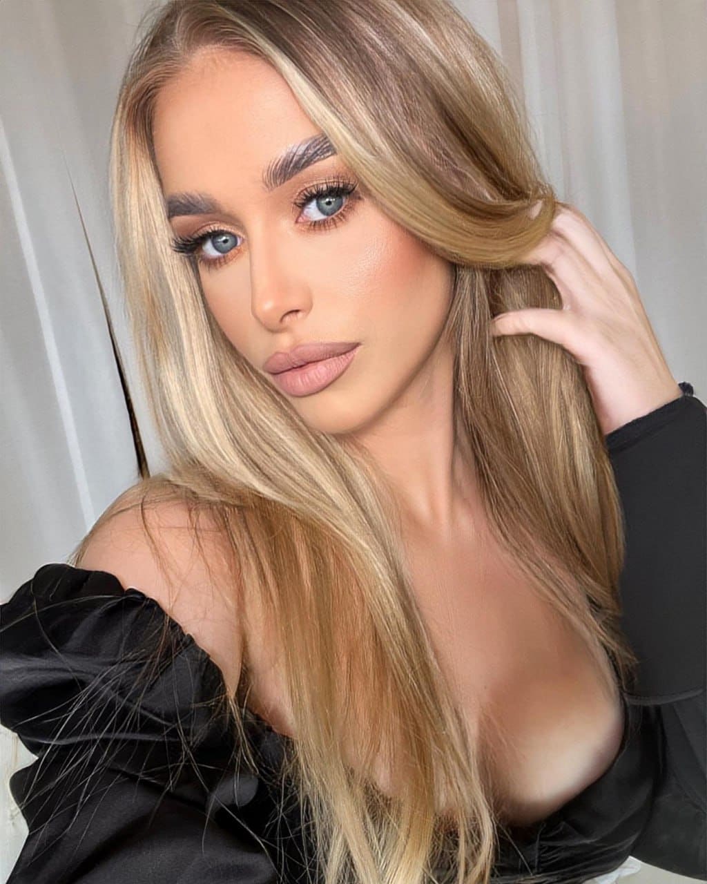 Sveta with long blonde hair in a black off-shoulder top, hand in hair, against a light curtain.