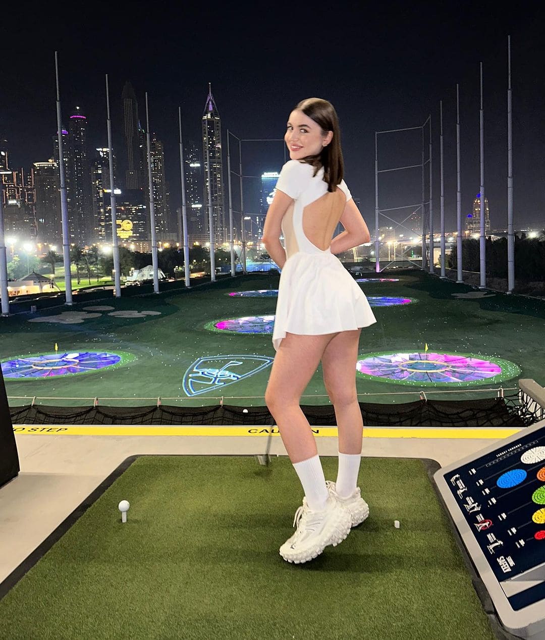 A woman stands on a rooftop golf range at night, wearing a white open-back dress, knee-high socks, and sneakers, with a city skyline in the background.