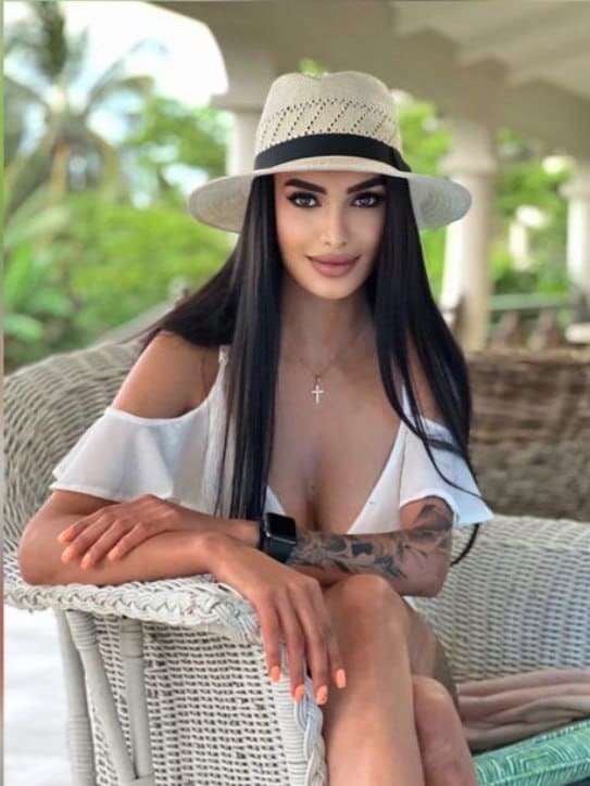 A woman with long dark hair sits on a wicker chair outdoors, wearing a white off-shoulder top, straw hat, and cross necklace, with a tattooed arm visible.