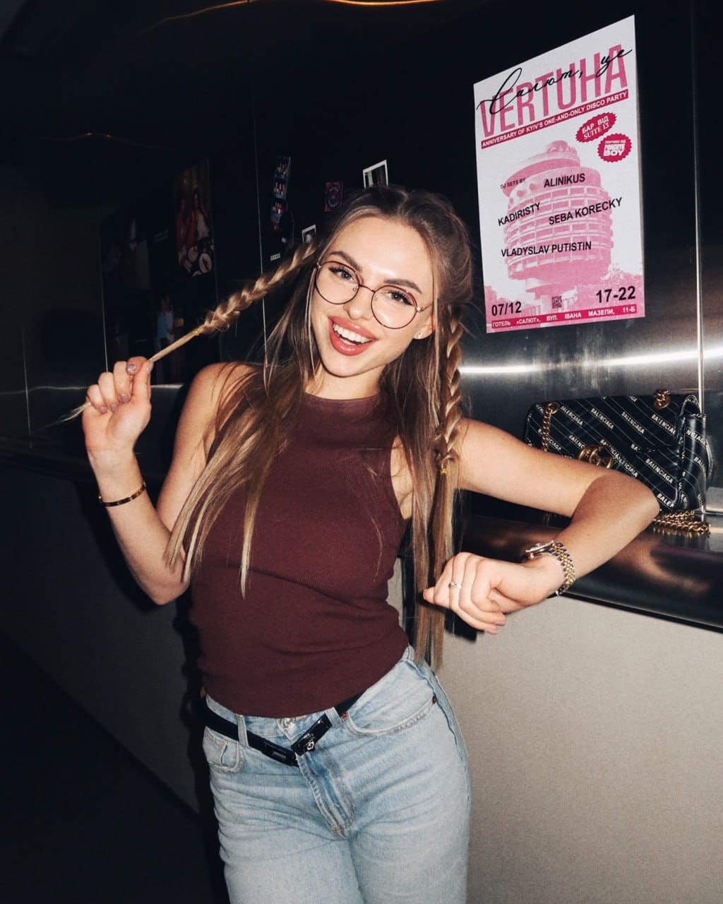 A blonde woman with braids and glasses smiles while leaning on a counter in a brown top and jeans, near a pink poster.