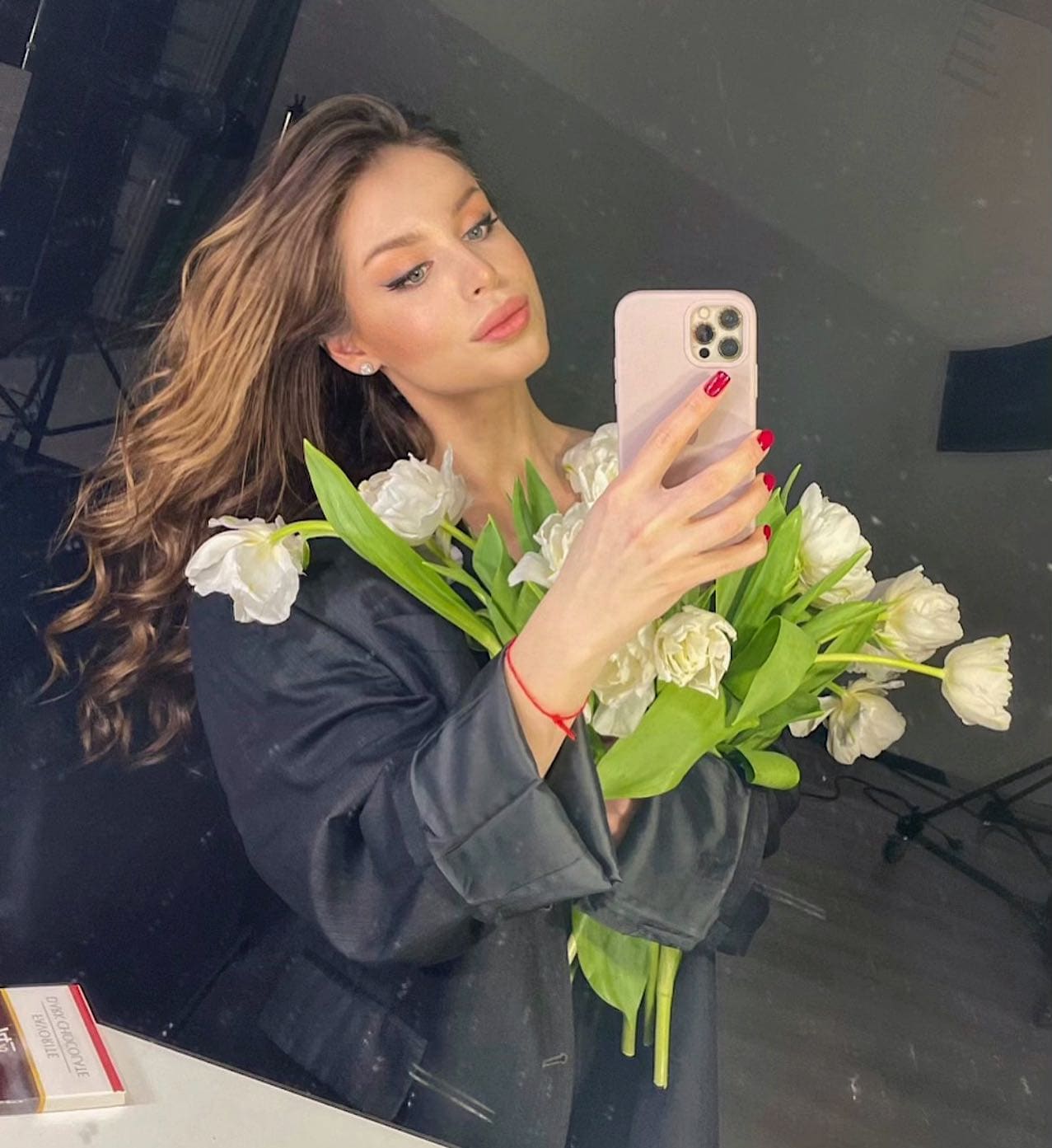 Mirror selfie holding white tulips, wearing a dark blazer and red nail polish in a dimly lit room.