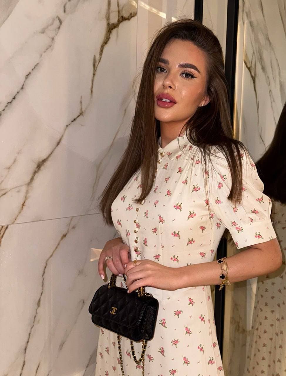Emilia stands before a marble wall in a white floral dress with a buttoned front, holding a black quilted handbag with a chain strap.