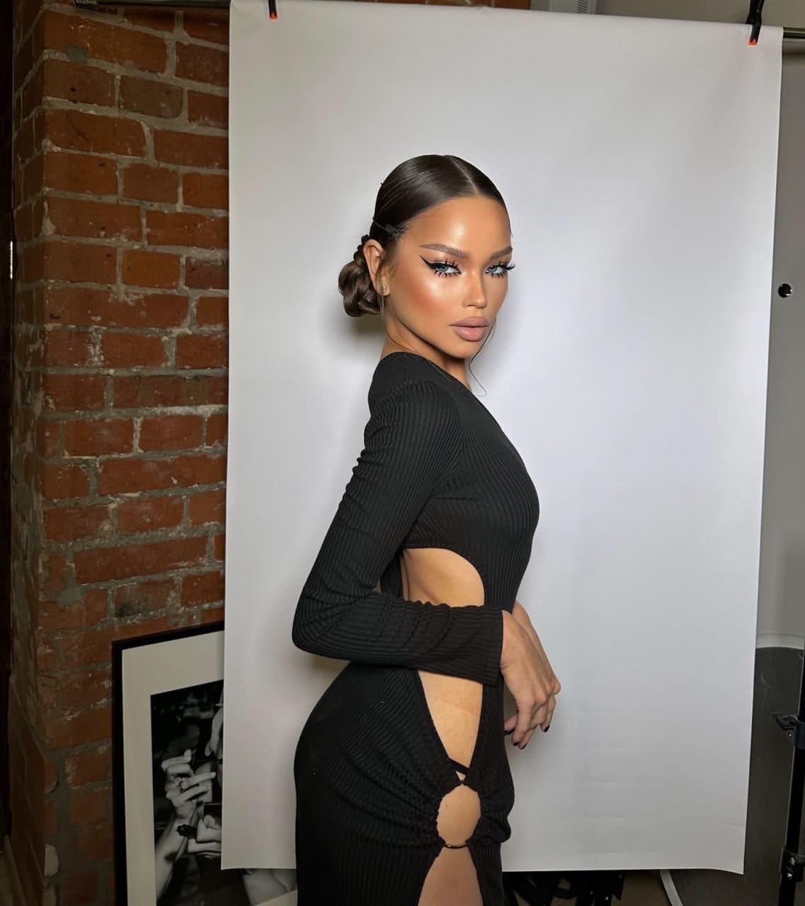 A woman in a black ribbed dress with cutouts stands in profile against a white backdrop near a brick wall.