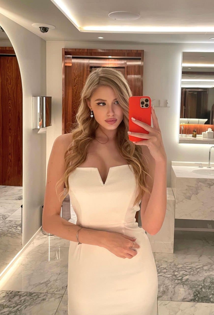 A woman takes a mirror selfie in a marble-floored bathroom, holding a red phone while wearing a strapless cream dress.
