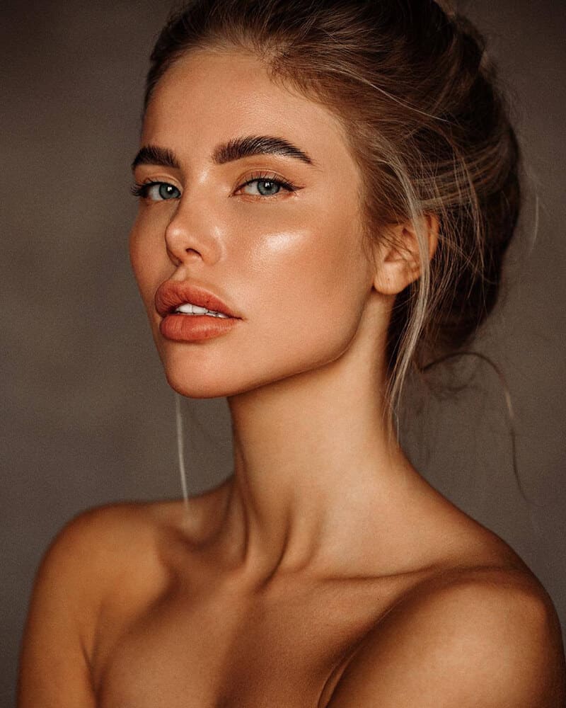 Close-up portrait of a woman with light brown hair in a loose bun, bare shoulders visible, set against a plain dark brown background.