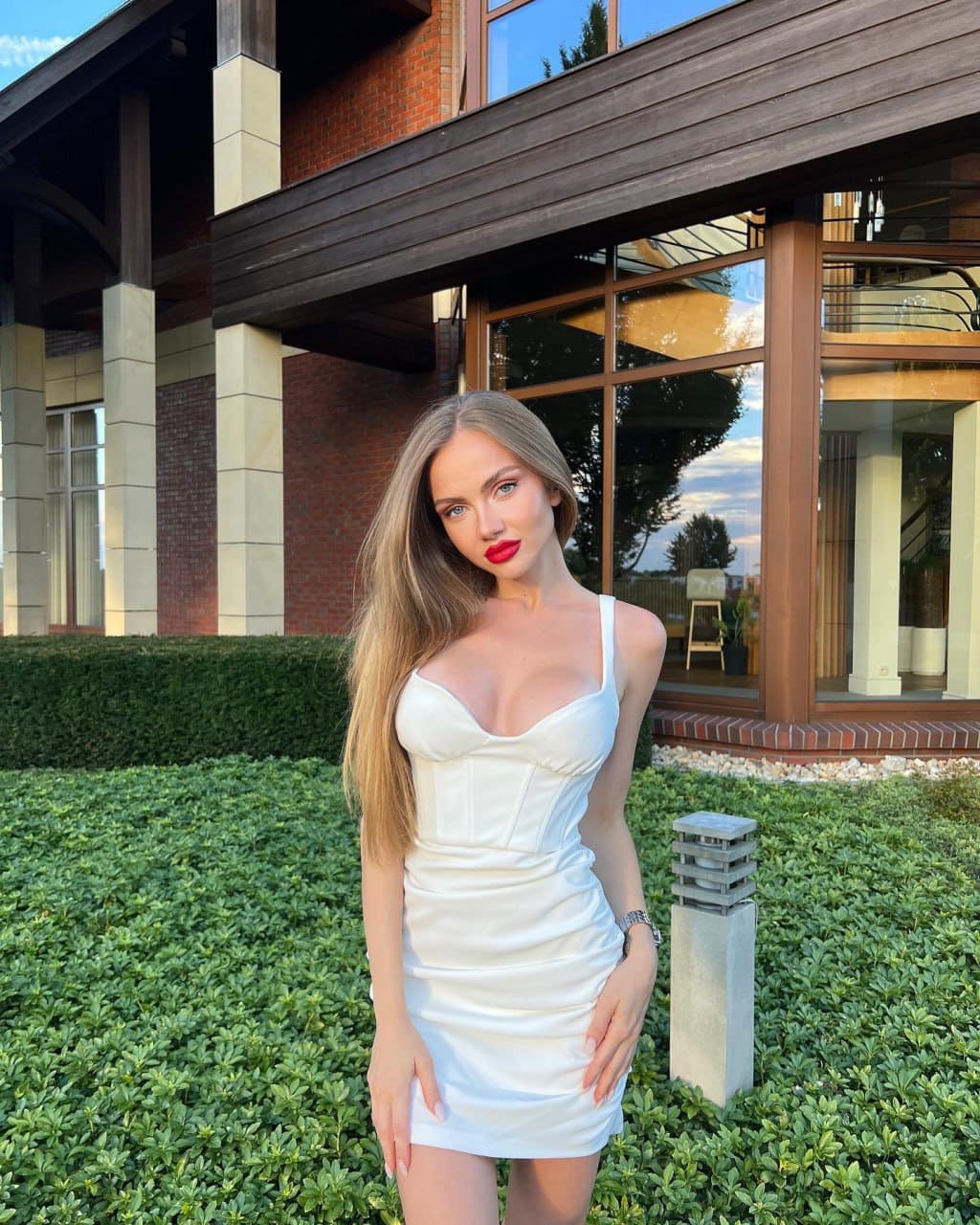 Selena stands in a garden before a brick building, wearing a white sleeveless mini dress and a watch on her left wrist.