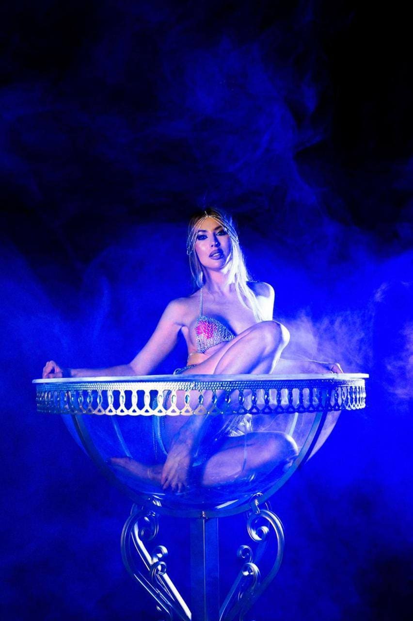 A blonde woman in a jeweled bikini top sits inside a large glass bowl on a stand, surrounded by blue stage lighting and smoke.