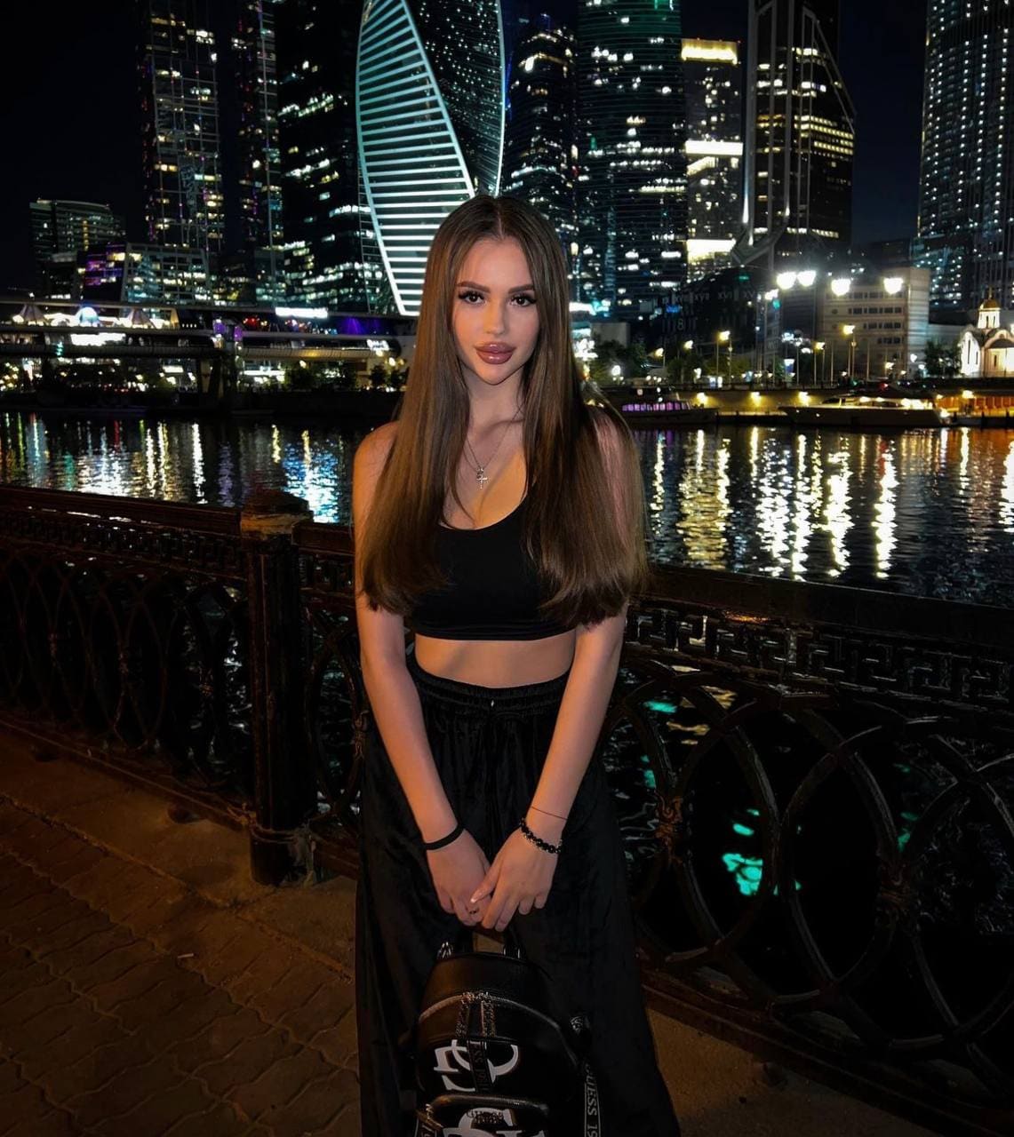 Tasya stands facing the camera in a black cropped tank top and wide-leg pants, holding a handbag against a night city skyline.
