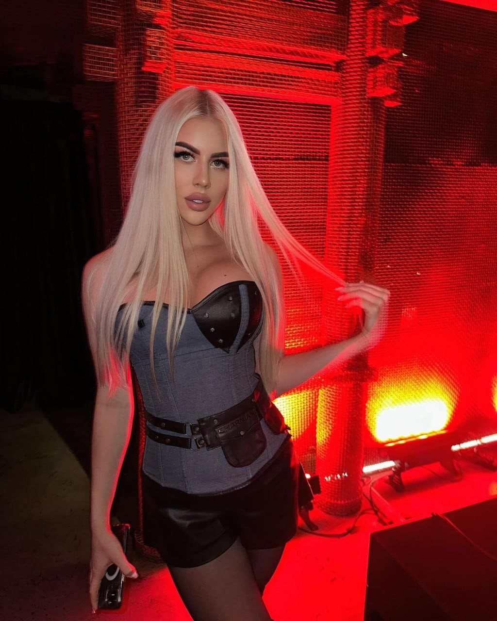 A woman stands in a red-lit industrial space wearing a corset and leather shorts, holding a camera and touching her hair.