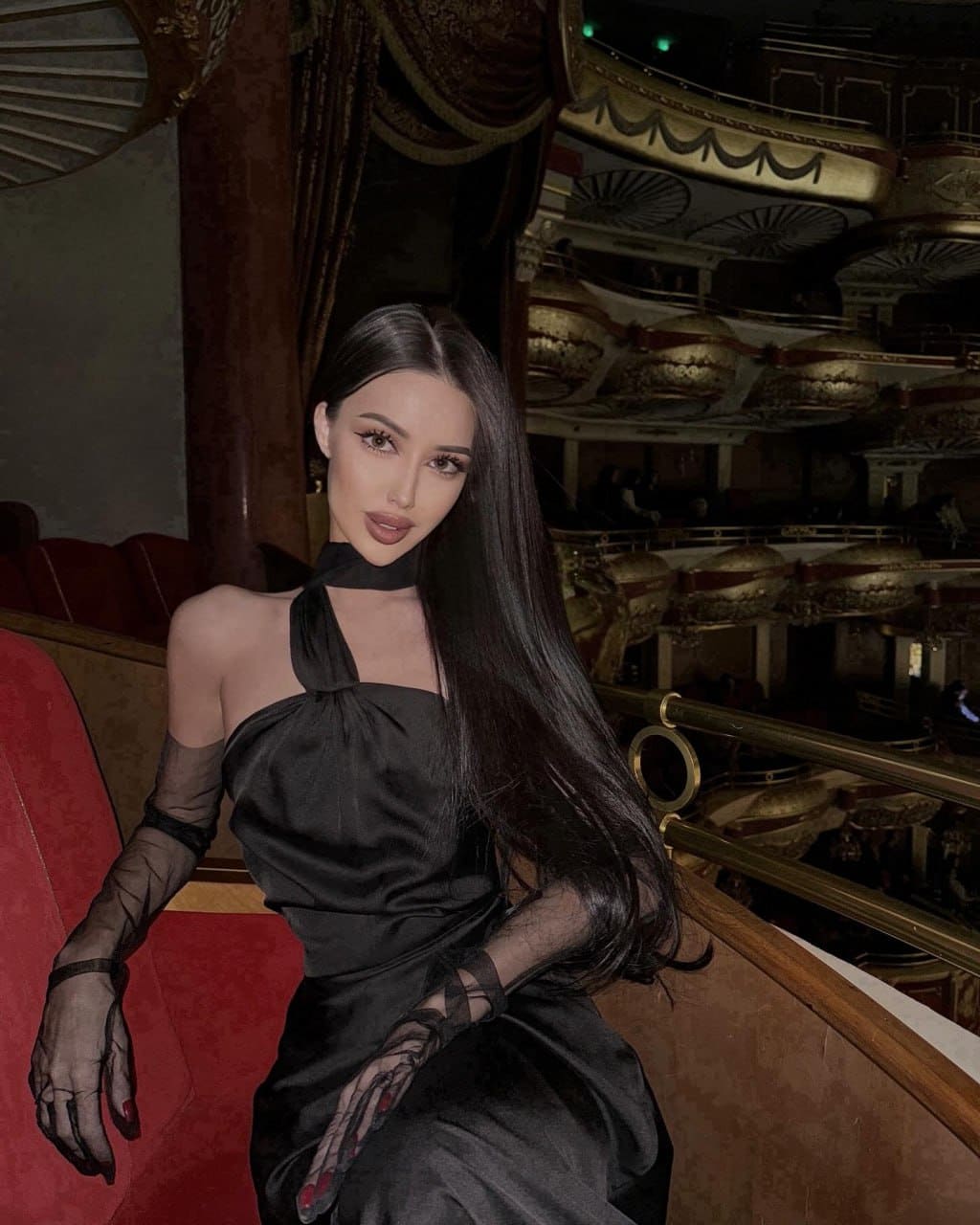 Seated in a red velvet theater seat, she wears a black halter dress with sheer sleeves and a matching neck tie.