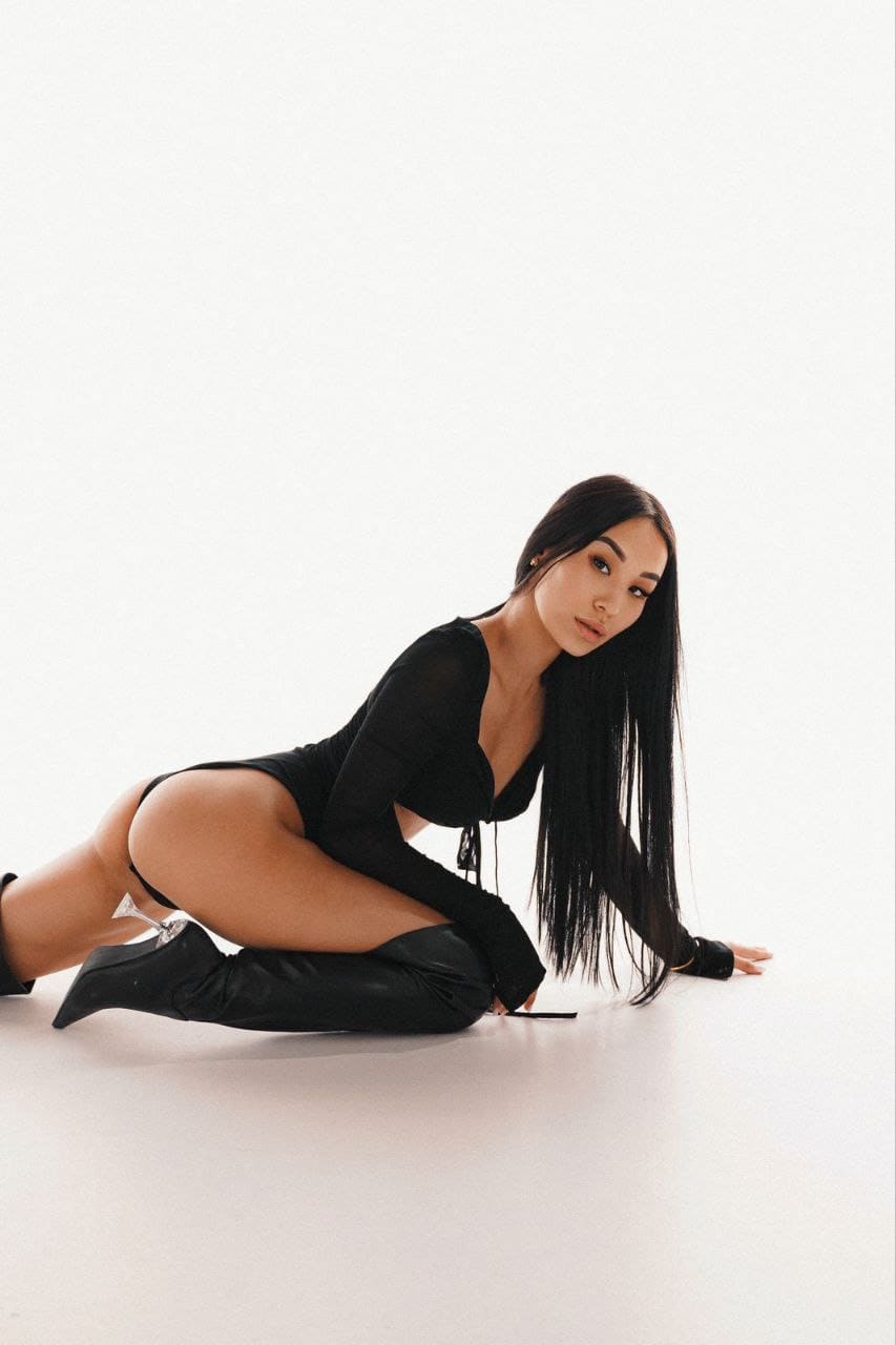 A woman kneels on a white floor in a black long-sleeved bodysuit and thigh-high boots, set against a plain white background.