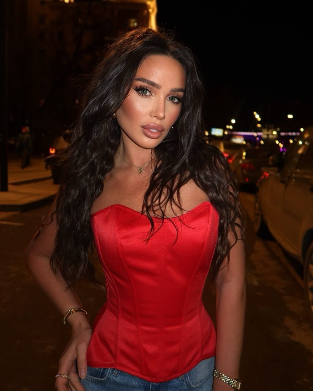 She stands on a city street at night in a red strapless satin top and blue jeans, with one hand resting on her hip.