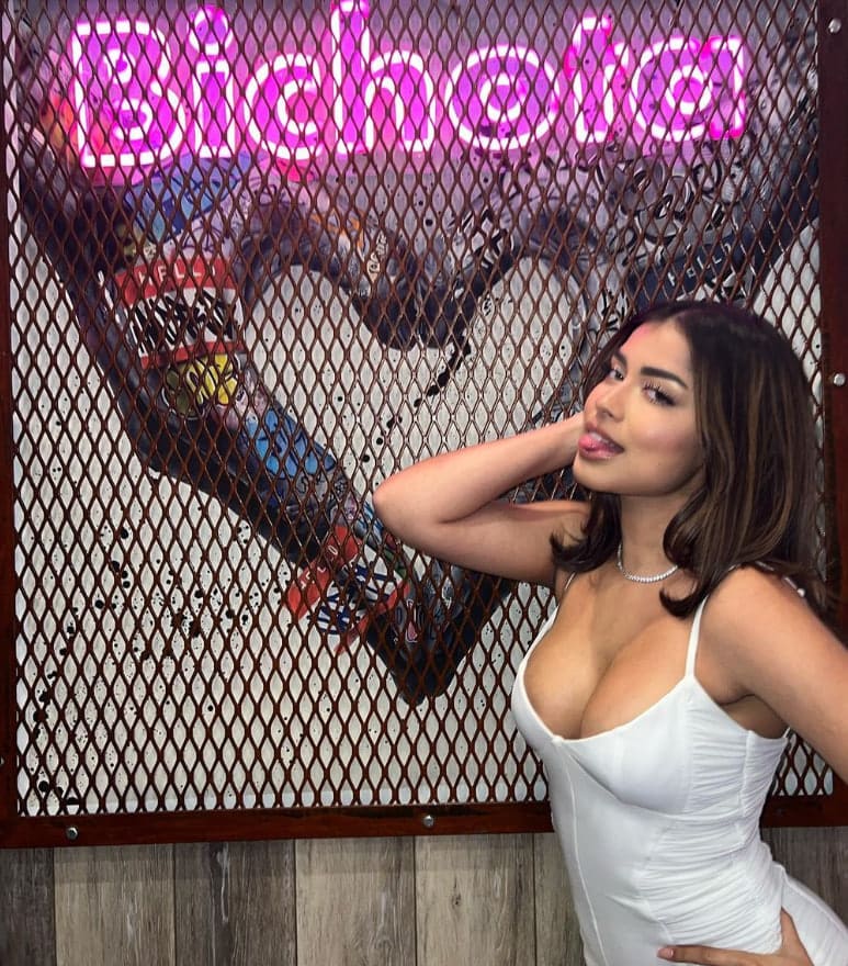 A woman with shoulder-length dark hair wears a white sleeveless dress, posing before a metal mesh screen featuring a pink neon sign and graffiti.