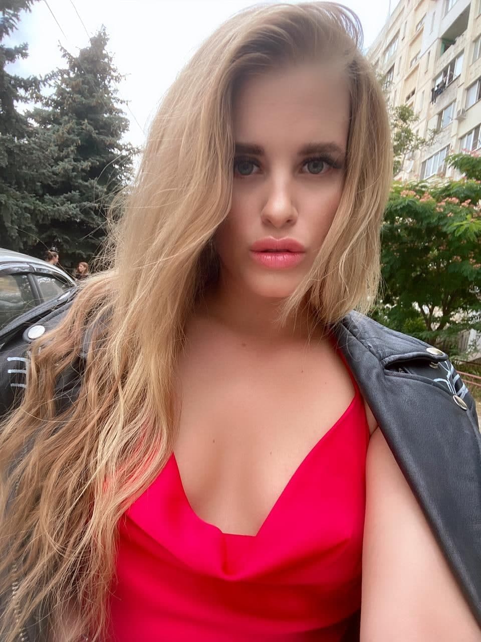 Marina outdoors in a red V-neck top and leather jacket, standing near trees and a building.
