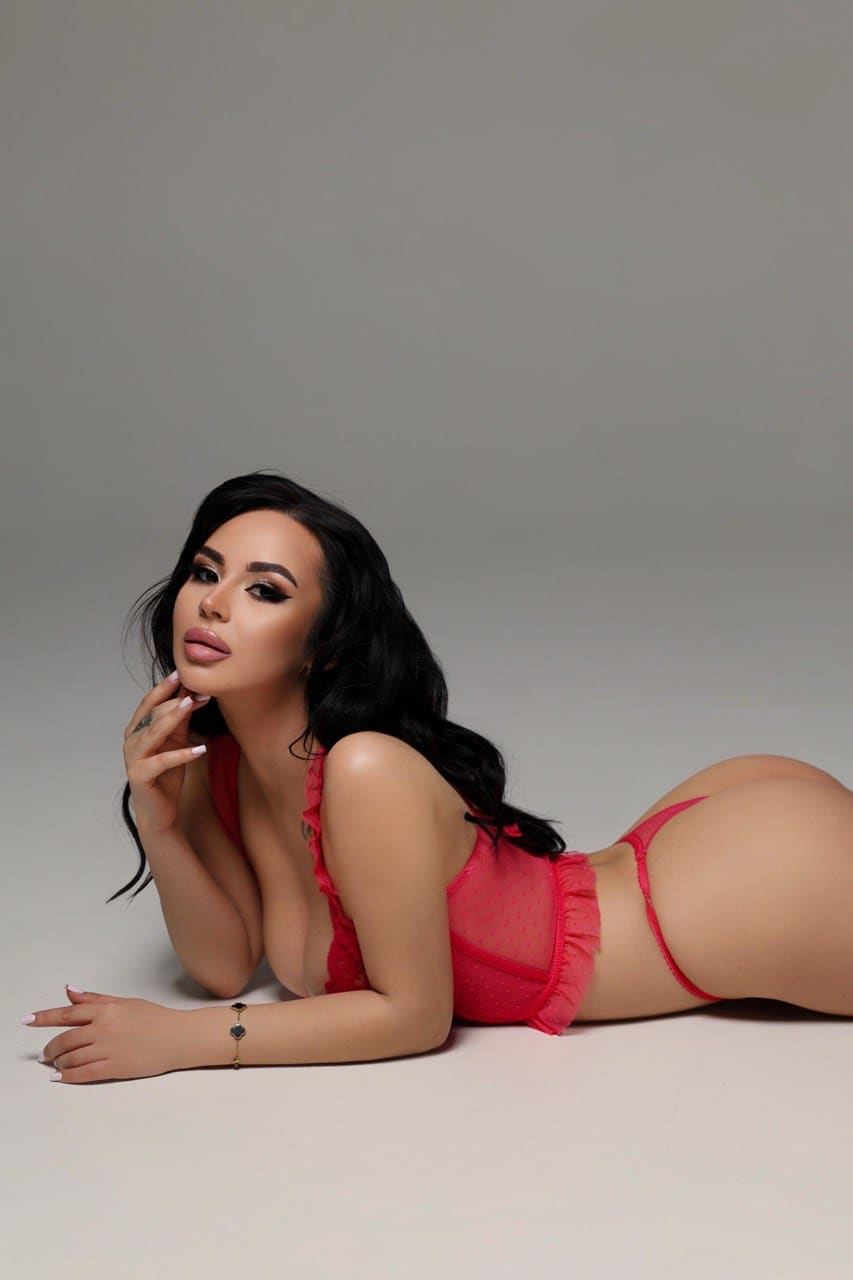 A woman lies on her side on a gray floor wearing a red ruffled lingerie set, with one hand near her chin.