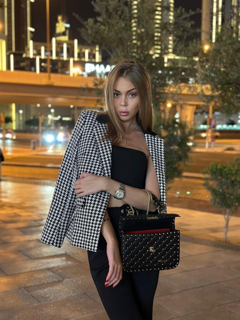 Woman in black strapless top and houndstooth blazer stands on a city sidewalk at night holding a handbag.