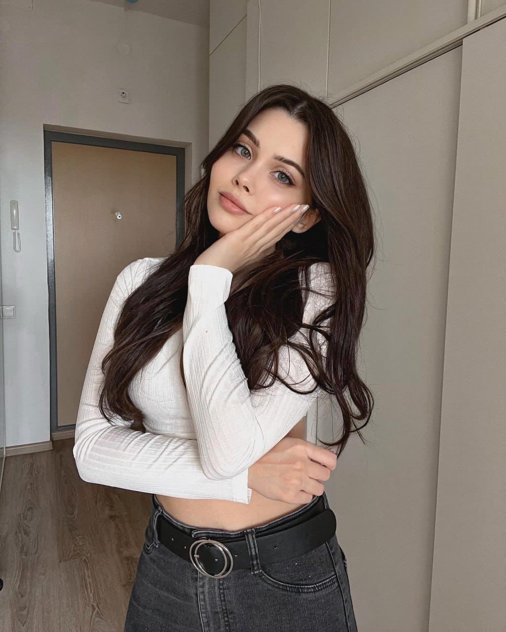 A woman in a white ribbed crop top and dark jeans stands indoors near a door, resting her right hand on her cheek.