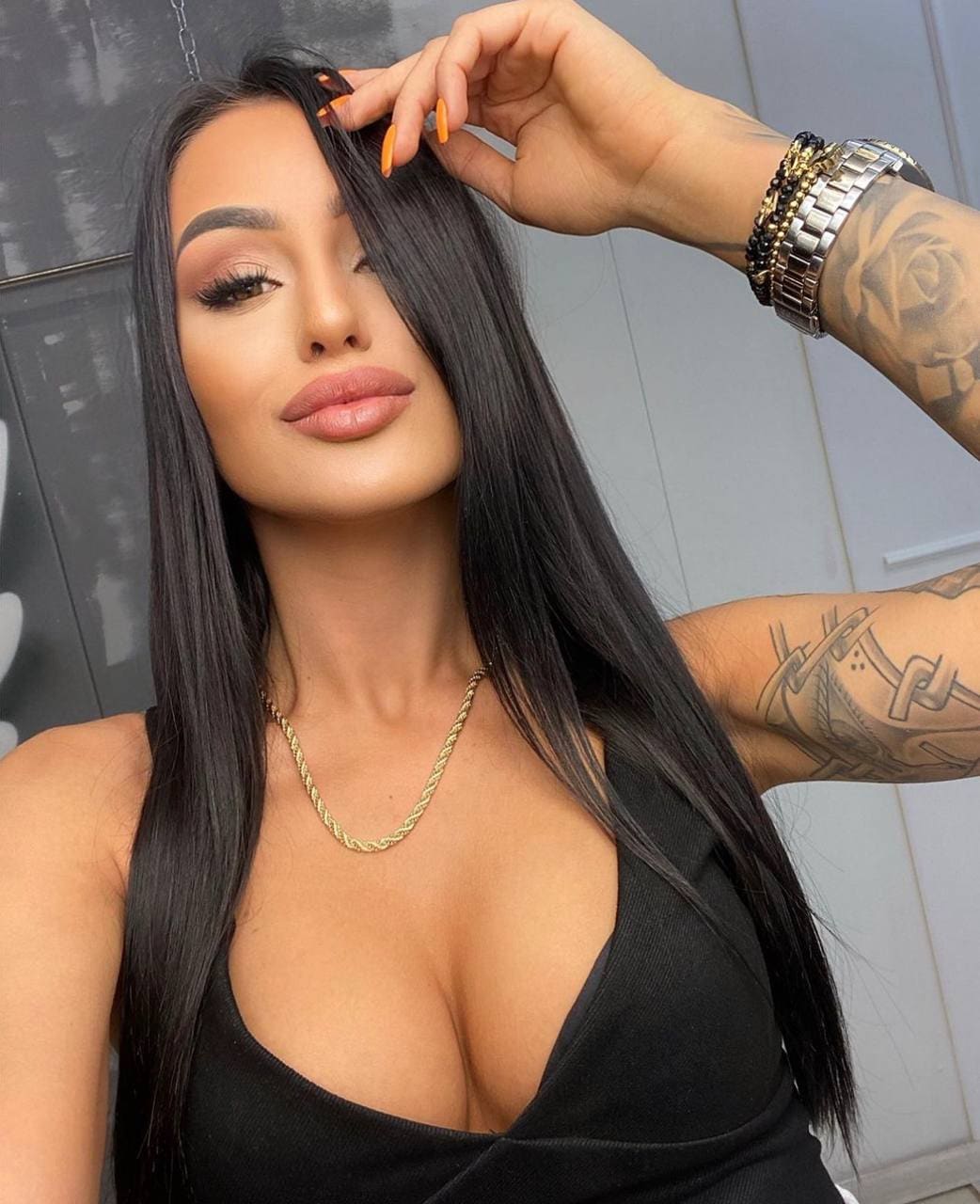 A woman with long dark hair and arm tattoos wears a black sleeveless top, gold chain necklace, and bracelets, with one hand raised to her hair.