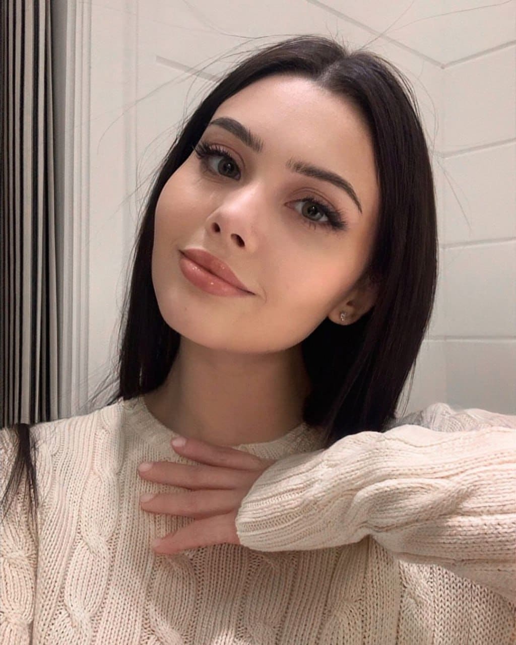 A woman in a cream cable-knit sweater rests a hand on her chest against a white paneled wall with a curtain in the background.