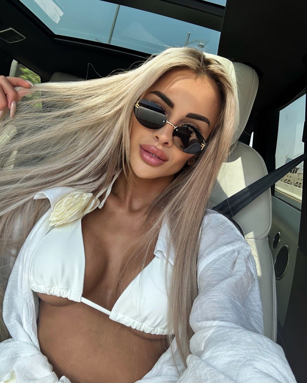A woman in dark sunglasses, a white bikini top with a flower accent, and an open white shirt sits in the back seat of a car.