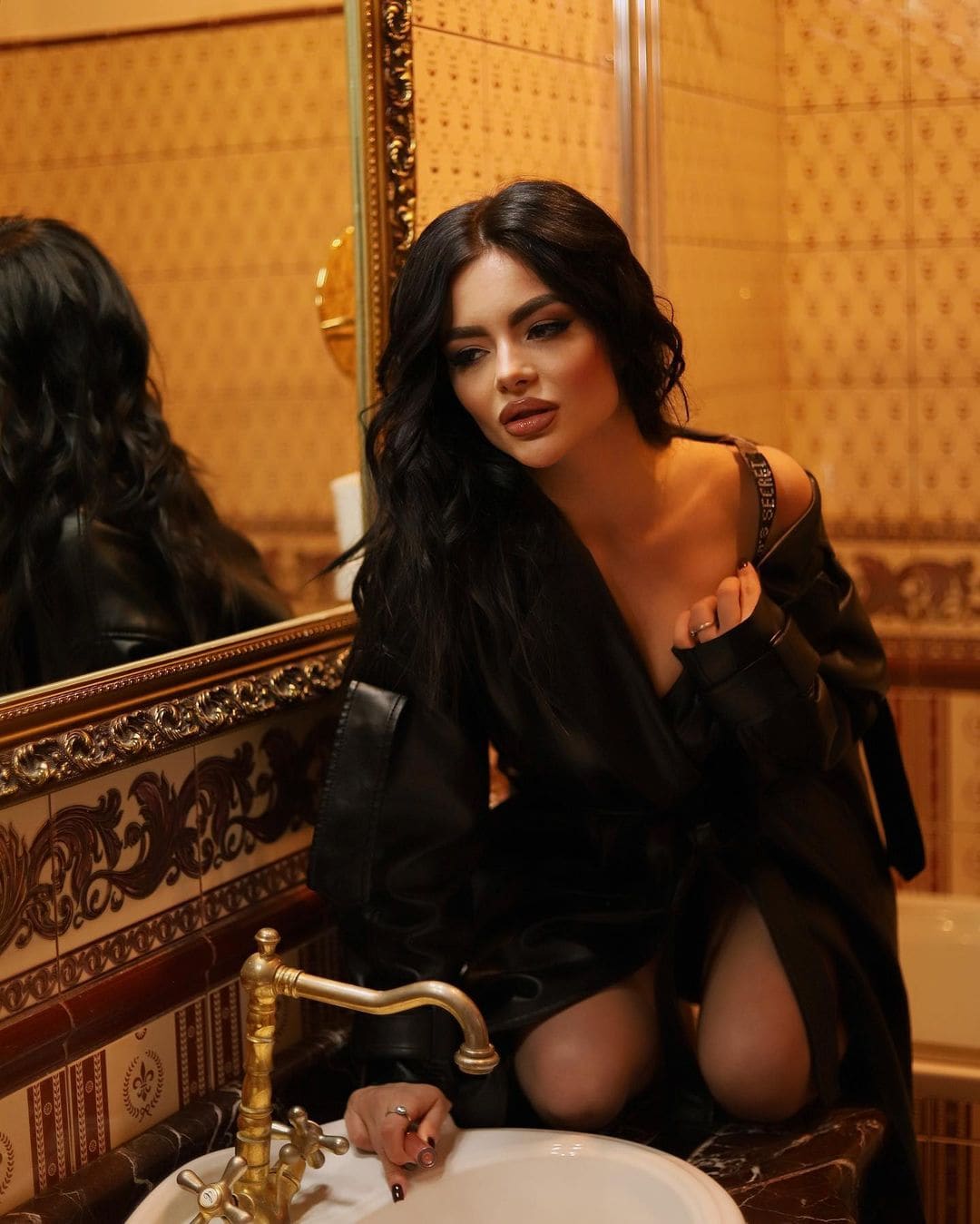 Kneeling on a marble counter, a woman in a black leather jacket and dress sits before a gold-framed mirror.