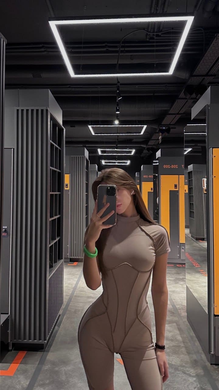 A woman takes a mirror selfie in a warehouse aisle wearing a beige high-neck bodysuit, holding a phone.