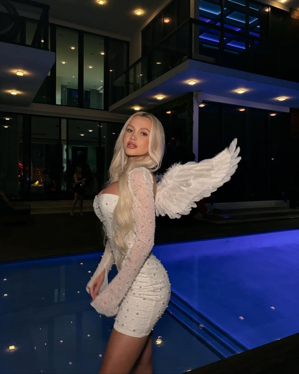 Inga stands by a blue-lit pool at night in a short white beaded dress with sheer sleeves and feathered wings, one hand on her hip.