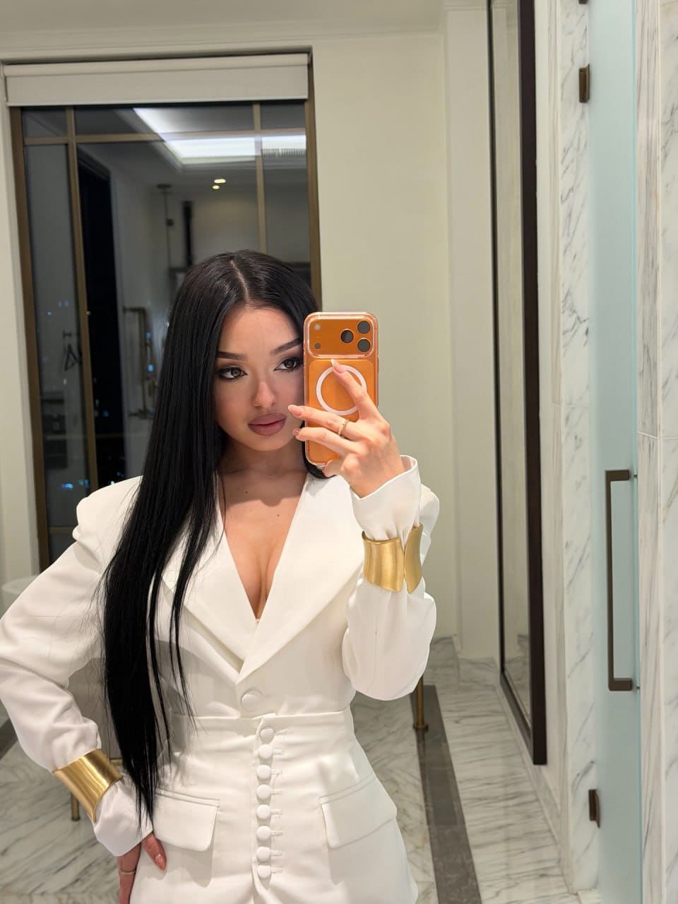 Mirror selfie of a woman in a white blazer with deep V-neck and gold bracelets, standing in a marble-tiled interior.
