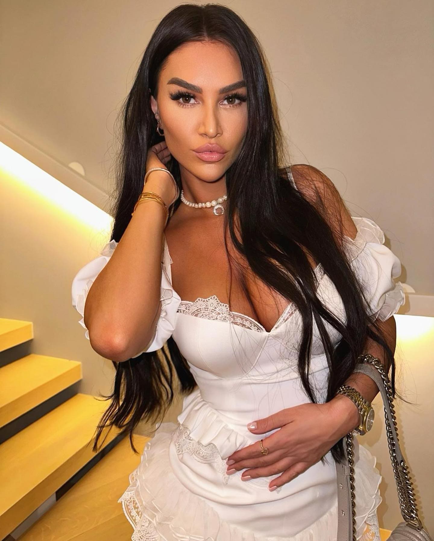 Dilya stands near an indoor staircase in a white off-shoulder lace dress, wearing a pearl necklace and holding a chain-strap bag.