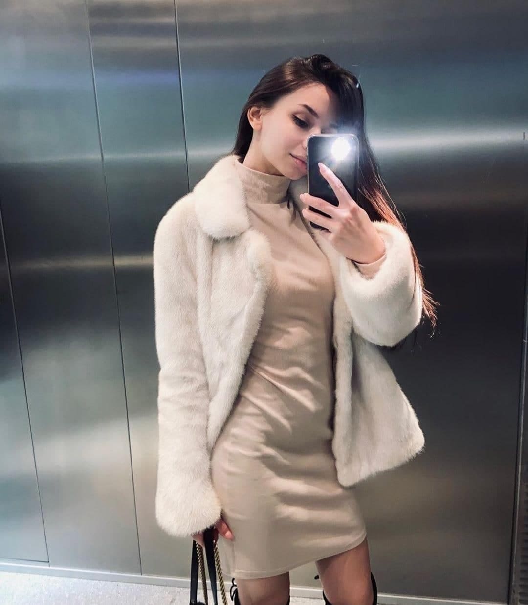 Zarina takes a mirror selfie in an elevator, wearing a beige turtleneck dress under a white faux-fur coat, holding a phone with the flash on.