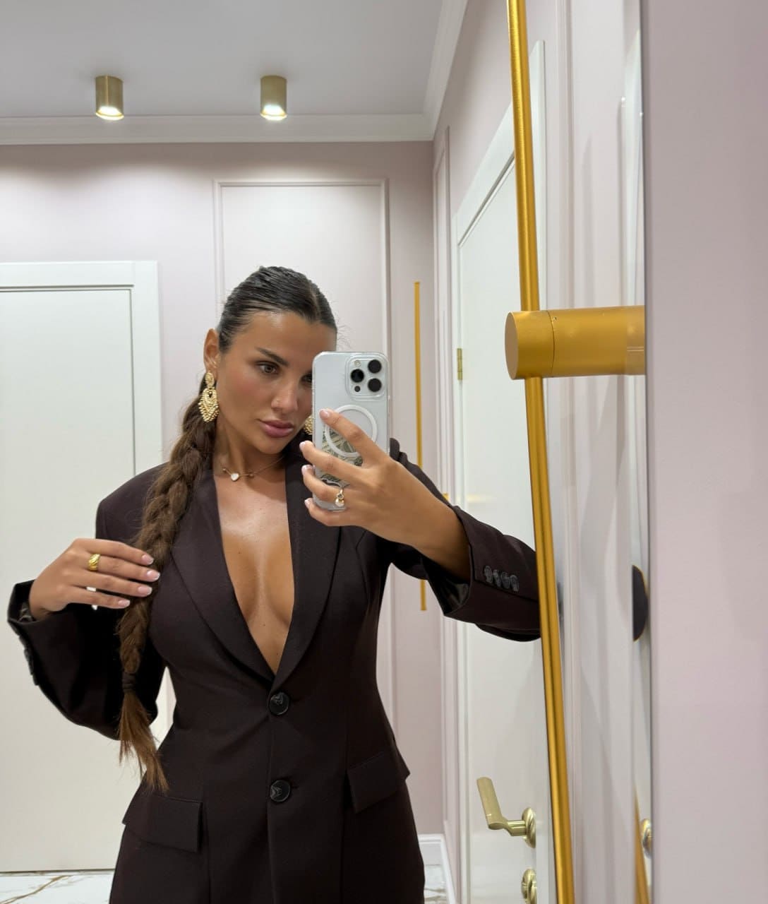 A woman with a dark braid takes a mirror selfie wearing a dark brown blazer with a deep V-neck, touching her hair in a pink room.
