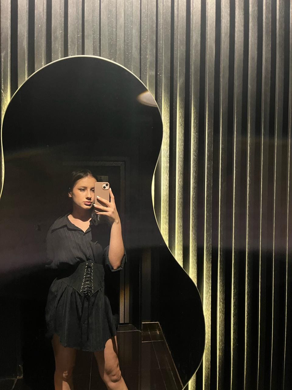 Selfie in an irregular mirror showing a woman in a black dress with lace-up bodice and puffed sleeves, against metallic slats.