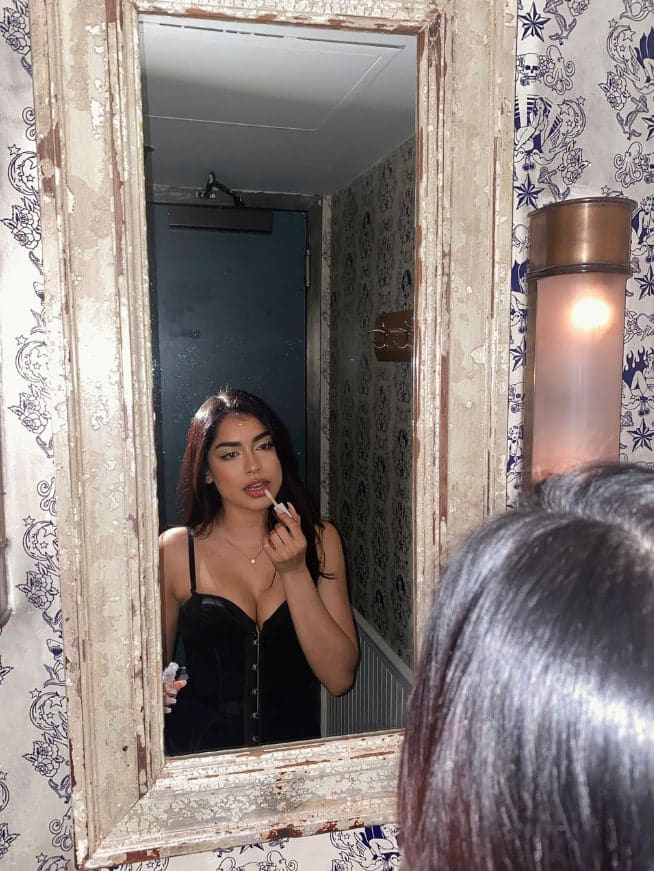 Seen in a mirror reflection, a woman applies lip product while wearing a black sleeveless top, set against patterned wallpaper and a wooden frame.