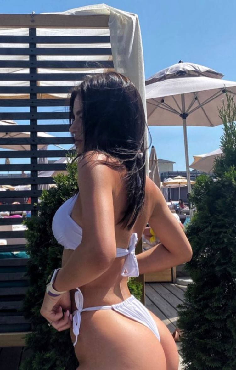 Standing in profile on a sunny deck, a woman wears a white string bikini with umbrellas in the background.