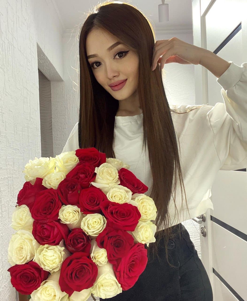 Vera stands indoors holding red and white roses, wearing a white long-sleeved top and dark pants, touching her hair.