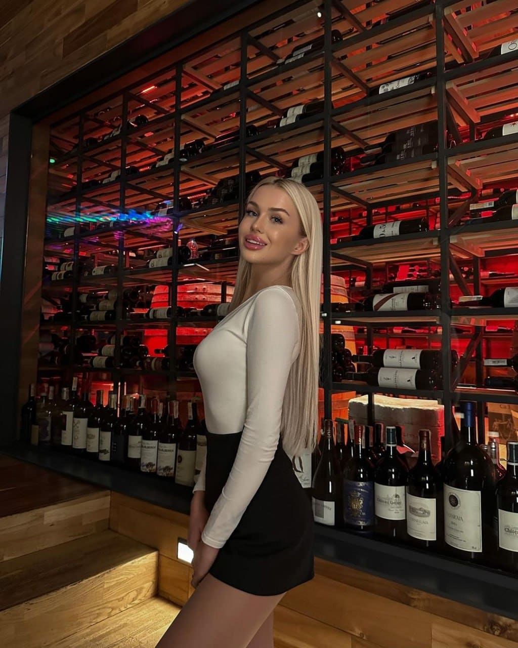 A blonde woman stands in profile before wine racks, wearing a white top and short black skirt.